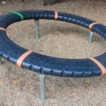 Ein kreisförmiger Spielplatz mit schwarzer, strukturierter Oberfläche, orangefarbenen Trennlinien und einem grünen Abschnitt, der von Metallfüßen getragen wird und auf einem Kiesboden steht.