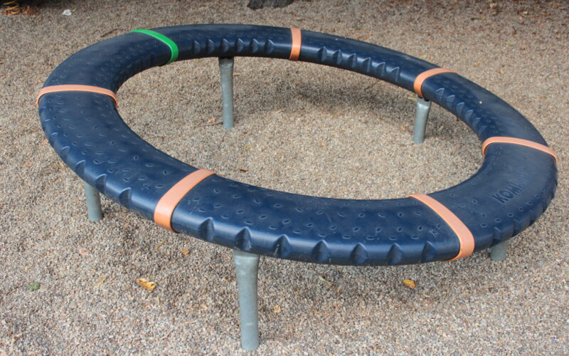 Ein kreisförmiger Spielplatz mit schwarzer, strukturierter Oberfläche, orangefarbenen Trennlinien und einem grünen Abschnitt, der von Metallfüßen getragen wird und auf einem Kiesboden steht.