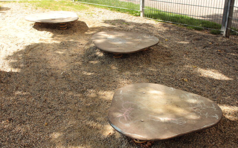 Drei flache, unregelmäßig geformte Metallplattformen mit Federn an der Unterseite befinden sich auf einer Kiesfläche in der Nähe eines Zauns, und auf den Plattformen sind einige Kreidezeichnungen zu sehen.