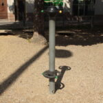 Eine Metallstange mit runden Griffen und einer runden Kappe steht auf einer Kiesfläche eines Spielplatzes und wirft einen Schatten auf den Boden.