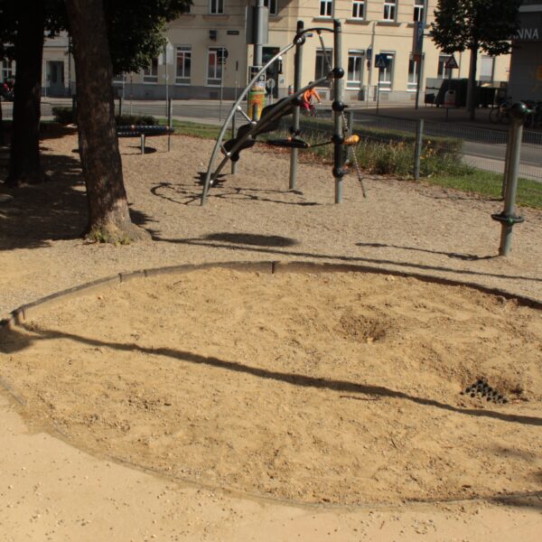 Ein kreisrunder Sandkasten mit aufgewühltem Sand befindet sich in einem schattigen Spielplatzbereich, im Hintergrund sind Klettergeräte und Gebäude zu sehen.