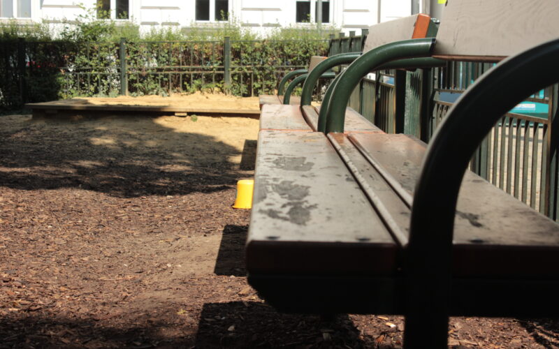 Eine hölzerne Parkbank mit grünen Metallarmlehnen vor einem eingezäunten Sandkastenbereich, mit Sonnenlicht und Schatten auf dem Boden.