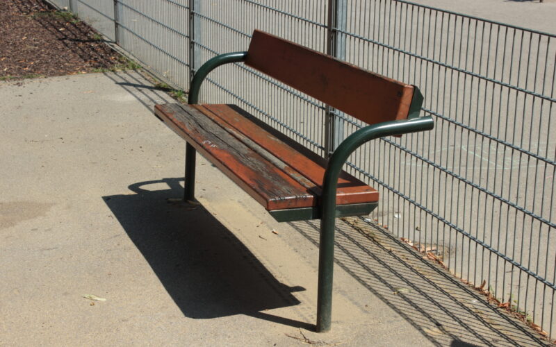 Eine abgenutzte Holzbank mit Metallbeinen und Rückenlehne steht auf einer Betonfläche neben einem Metallzaun in einem Außenbereich.
