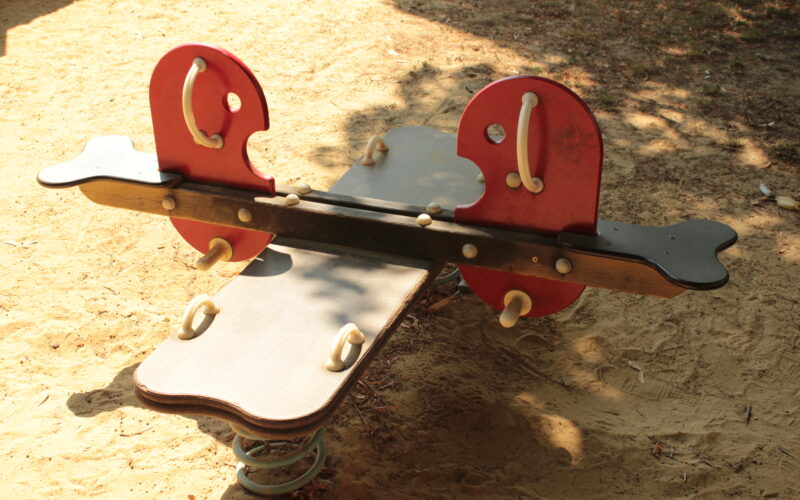 Eine Spielplatzwippe mit roten Sitzen in Form von Fischen steht auf sandigem Boden im Halbschatten.