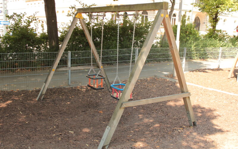 Holzschaukel mit zwei Schalensitzen auf einem mit Mulch bedeckten Spielplatz, umgeben von einem Metallzaun und Bäumen.