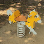 Ein gelbes und weißes gefedertes Schaukelpferd steht auf sandigem Boden im Halbschatten.