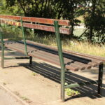 Eine abgenutzte Parkbank aus Holz mit Metallrahmen steht auf einem gepflasterten Weg neben Gras und Bäumen und wird vom Sonnenlicht beschattet.