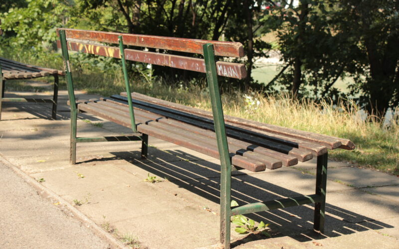 Eine abgenutzte Parkbank aus Holz mit Metallrahmen steht auf einem gepflasterten Weg neben Gras und Bäumen und wird vom Sonnenlicht beschattet.