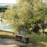 Eine grüne Bank steht an einem Uferweg, im Hintergrund eine Brücke und ein Boot auf dem Wasser. Bäume und Grün umgeben die Szene.