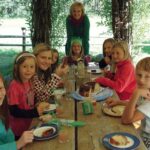 Eine Gruppe von Kindern und ein Erwachsener sitzen lächelnd an einem Holztisch im Freien und essen Kuchen, umgeben von Grün und natürlichem Licht.