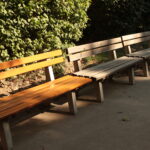 Entlang eines gepflasterten Weges in einem Park stehen mehrere leere Holzbänke, im Hintergrund grüne Sträucher und das Sonnenlicht wirft Schatten.