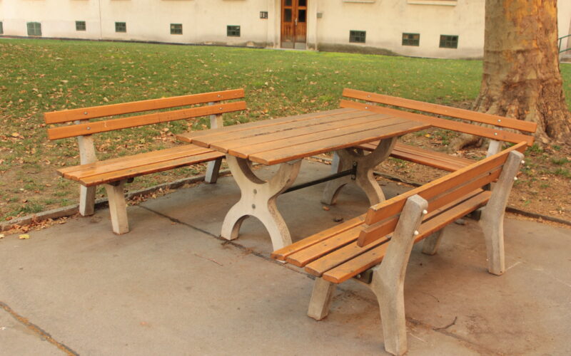 Picknicktisch aus Holz mit zwei Bänken und einer Sitzgelegenheit, aufgestellt auf einer Betonfläche neben einem Baum in einem Park im Freien.