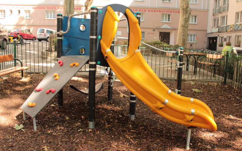 Ein Spielplatz mit einer gelben Rutsche, einer kleinen Kletterwand und einer blauen Platte, umgeben von Mulch und einem Metallzaun in einem städtischen Wohngebiet.