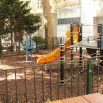 Ein kleiner eingezäunter Spielplatz mit einer gelben Rutsche, einem Klettergerüst und drehenden Spielgeräten, umgeben von Bäumen und Gebäuden.