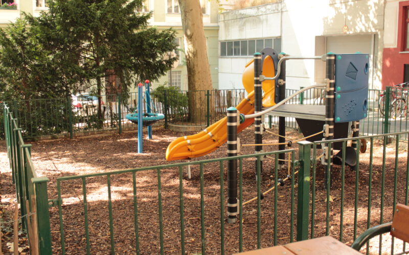 Ein kleiner eingezäunter Spielplatz mit einer gelben Rutsche, einem Klettergerüst und drehenden Spielgeräten, umgeben von Bäumen und Gebäuden.