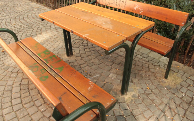 Ein Holztisch für den Außenbereich mit zwei Bänken auf einer Kopfsteinpflasterfläche. Die Bank und der Tisch haben grüne Metallbeine und einige Graffiti auf dem Holz.