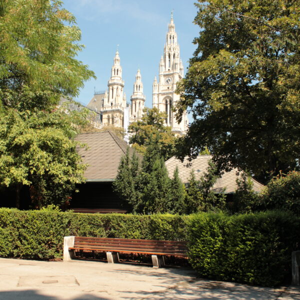Ein Park mit leeren Bänken, grünen Bäumen und den Türmen eines großen historischen Gebäudes, die im Hintergrund unter einem blauen Himmel sichtbar sind.