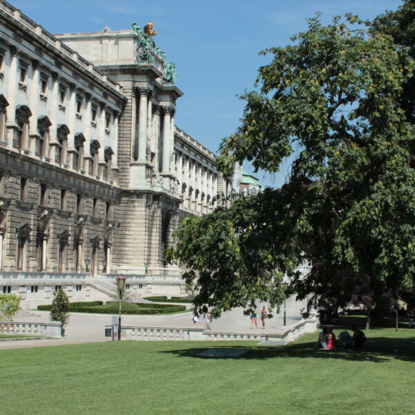 Ein großes historisches Gebäude mit kunstvoller Architektur steht neben einem grünen Rasen und Bäumen, und Menschen gehen unter einem klaren Himmel im Park spazieren und sitzen.