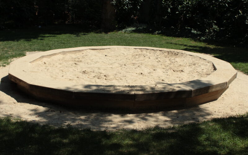 Ein leerer achteckiger Sandkasten mit Holzseiten steht auf Gras in einem schattigen Außenbereich.