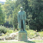 Eine Bronzestatue eines Mannes in langem Mantel und Hut steht auf einem Steinsockel, umgeben von Grün in einem Park.