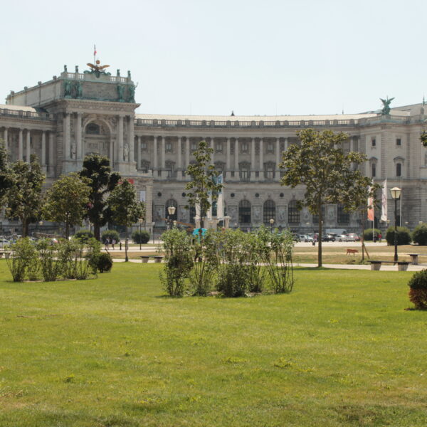 Neoklassizistisches Palastgebäude mit Statuen und Säulen, an einem sonnigen Tag von einem grasbewachsenen Park mit Bäumen und Sträuchern im Vordergrund aus gesehen.