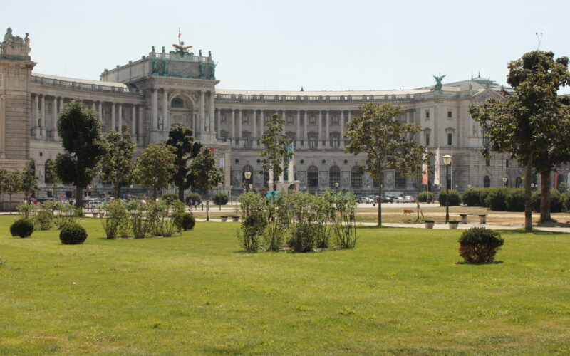 Neoklassizistisches Palastgebäude mit Statuen und Säulen, an einem sonnigen Tag von einem grasbewachsenen Park mit Bäumen und Sträuchern im Vordergrund aus gesehen.