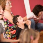 Eine Gruppe von Erwachsenen und Kindern sitzt oder hockt drinnen und lacht und lächelt gemeinsam vor einer roten Wand.