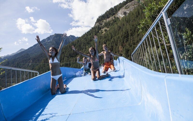 Vier Kinder rutschen im Freien, umgeben von Bäumen und Bergen unter einem teilweise bewölkten Himmel, eine blaue Wasserrutsche hinunter.