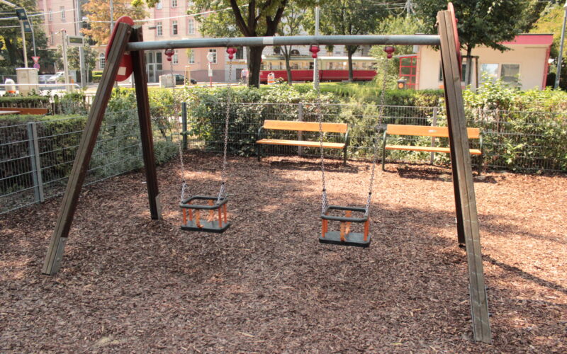 Zwei leere Schaukeln für Kleinkinder hängen an einem Holzrahmen auf einem Spielplatz, mit Bänken, Zäunen und Grünpflanzen im Hintergrund.