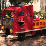 Ein rot-gelbes Holzspielgerät in Form eines Lastwagens steht auf Mulch in einem Park mit Bäumen und Bänken.