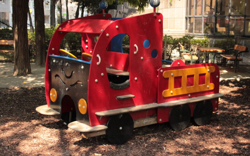 Ein rot-gelbes Holzspielgerät in Form eines Lastwagens steht auf Mulch in einem Park mit Bäumen und Bänken.