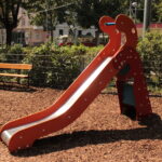 Eine kleine rote Kinderrutsche mit einer Metalloberfläche steht auf Holzspänen auf einem Spielplatz, mit Bänken und Grünpflanzen im Hintergrund.