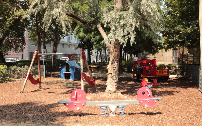 Ein Spielplatz mit einer Wippe, Schaukeln, einer Rutsche und einem roten Spielzeug-Feuerwehrauto, umgeben von Bäumen und Holzspänen.