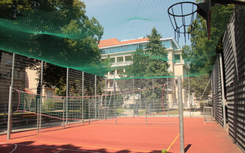 Ein roter Sportplatz mit Basketballkorb, Badmintonnetz und grünem Sicherheitsnetz, umgeben von einem Metallzaun und Bäumen.