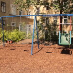 Ein Spielplatz mit blauen Metallschaukeln, darunter ein grüner barrierefreier Schaukelsitz, steht auf einer Mulchfläche in der Nähe von Sträuchern und einem alten Wohnhaus.