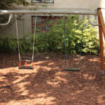 Eine Schaukel aus Metall und Holz mit zwei leeren Schaukeln steht auf einem mit Holzspänen bedeckten Spielplatz, mit Grünpflanzen und einem Gebäude im Hintergrund.
