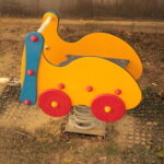 Ein gelber und roter Springreiter in Form eines Autos ist auf dem Sandboden eines Spielplatzes installiert.