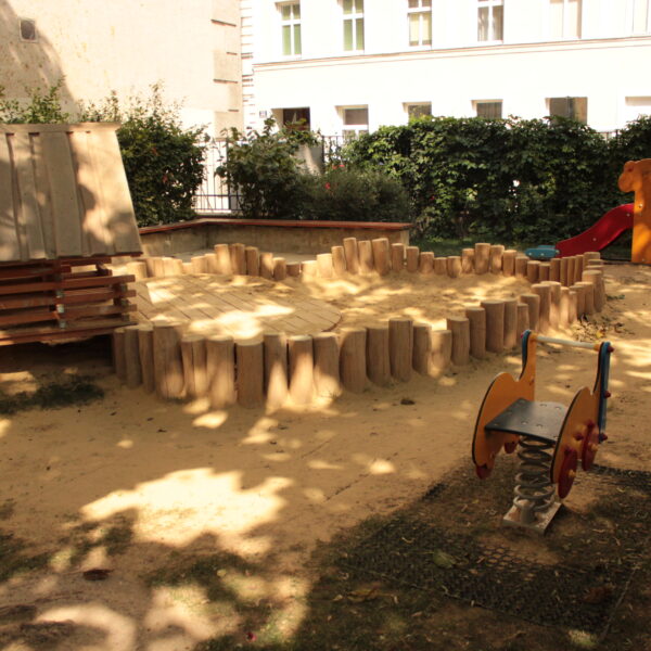 Ein kleiner Spielplatz mit einem Sandbereich, einem Holzspielgerät, einer orangefarbenen Rutsche und einem Springreiter, umgeben von Büschen und einem Gebäude im Hintergrund.