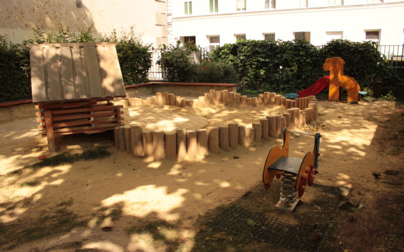 Ein kleiner Spielplatz mit einem Sandbereich, einem Holzspielgerät, einer orangefarbenen Rutsche und einem Springreiter, umgeben von Büschen und einem Gebäude im Hintergrund.