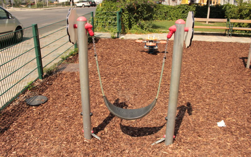 Ein flexibler Spielplatz-Schaukelsitz ist zwischen zwei Metallpfosten über einem Holzschnitzel-Bodenbelag aufgehängt; im Hintergrund sind ein Zaun und Bänke zu sehen.