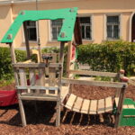 Spielplatz aus Holz mit einer Rutsche, einer Kletterbrücke und einem Aussichtsbereich, der auf Mulch liegt, mit einer Hecke und einem Gebäude im Hintergrund.