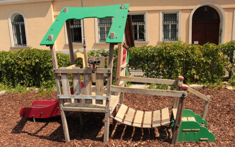 Spielplatz aus Holz mit einer Rutsche, einer Kletterbrücke und einem Aussichtsbereich, der auf Mulch liegt, mit einer Hecke und einem Gebäude im Hintergrund.