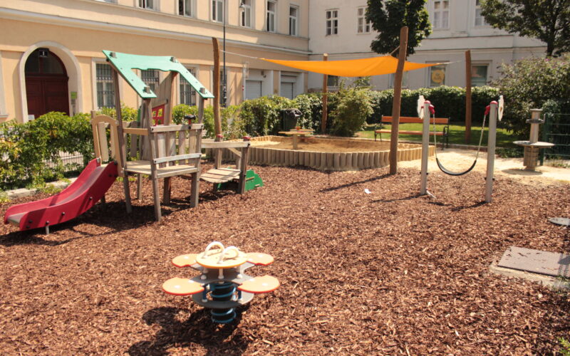 Kinderspielplatz mit Klettergerüst, Rutsche, Sandkasten mit Überdachung, Springreiter, Schaukel und Bänken, auf Mulch in einem von Gebäuden umgebenen Innenhof gelegen.