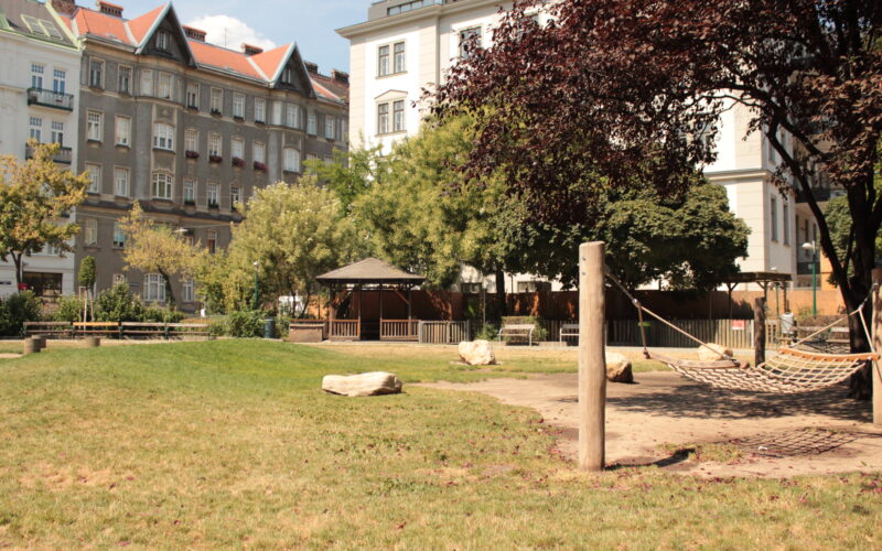 Ein Stadtpark mit Hängematte, grünem Rasen, großen Felsen, Bäumen und einem Pavillon, umgeben von mehrstöckigen Wohngebäuden.