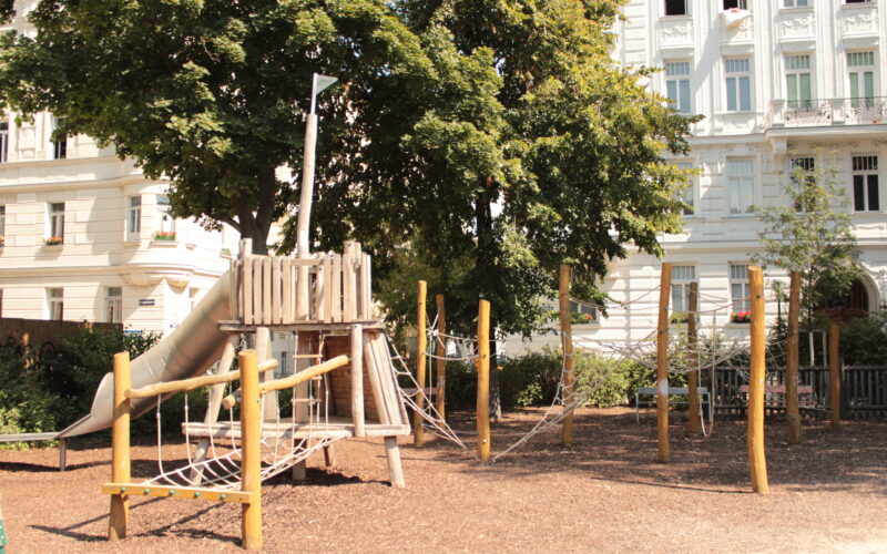 Ein Spielplatz aus Holz mit Seilbrücken und einer Rutsche befindet sich in einem Park, umgeben von Bäumen und angrenzend an weiße Wohnhäuser.