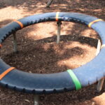 Ein großes kreisförmiges blaues Spielgerät mit orangefarbenen und grünen Teilen, das von Metallfüßen getragen wird, steht auf einem mit Rinde bedeckten Boden im gedämpften Sonnenlicht.