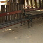 Zwei leere Holzbänke mit Metallarmlehnen stehen auf einem unbefestigten Boden in der Nähe eines Holzzauns in einem Außenbereich.