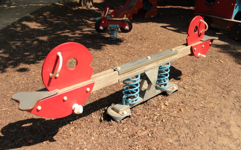 Rote und graue Wippe mit Federstützen auf einem Spielplatz mit Mulchbodenbelag, bei Tageslicht fotografiert.
