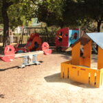 Ein Spielplatz mit einer Federwippe, einem Spielhaus und Klettergerüsten, umgeben von Bäumen und einem Holzzaun an einem sonnigen Tag.