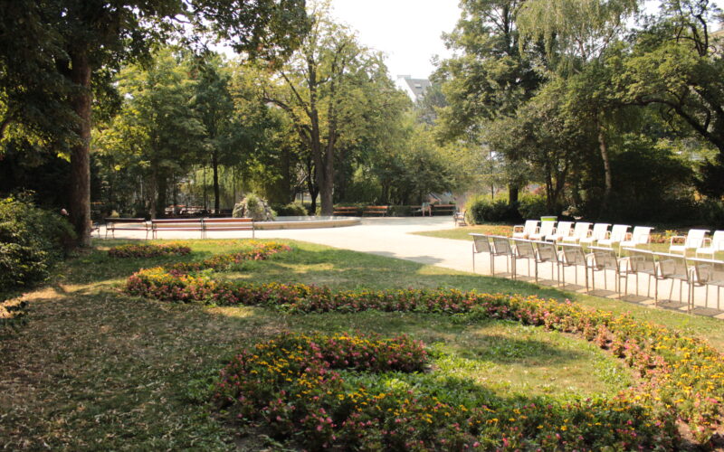 Eine angelegte Parkanlage mit geschwungenen Blumenbeeten, in Reihen angeordneten leeren weißen Stühlen und Bäumen, die an einem sonnigen Tag Schatten spenden.